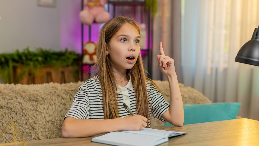 Child girl sitting at home table does homework with notebook open thoughtfully looking for problems solution. School kid happily finds correct answer, excitedly smiling, exclaiming joyful understand