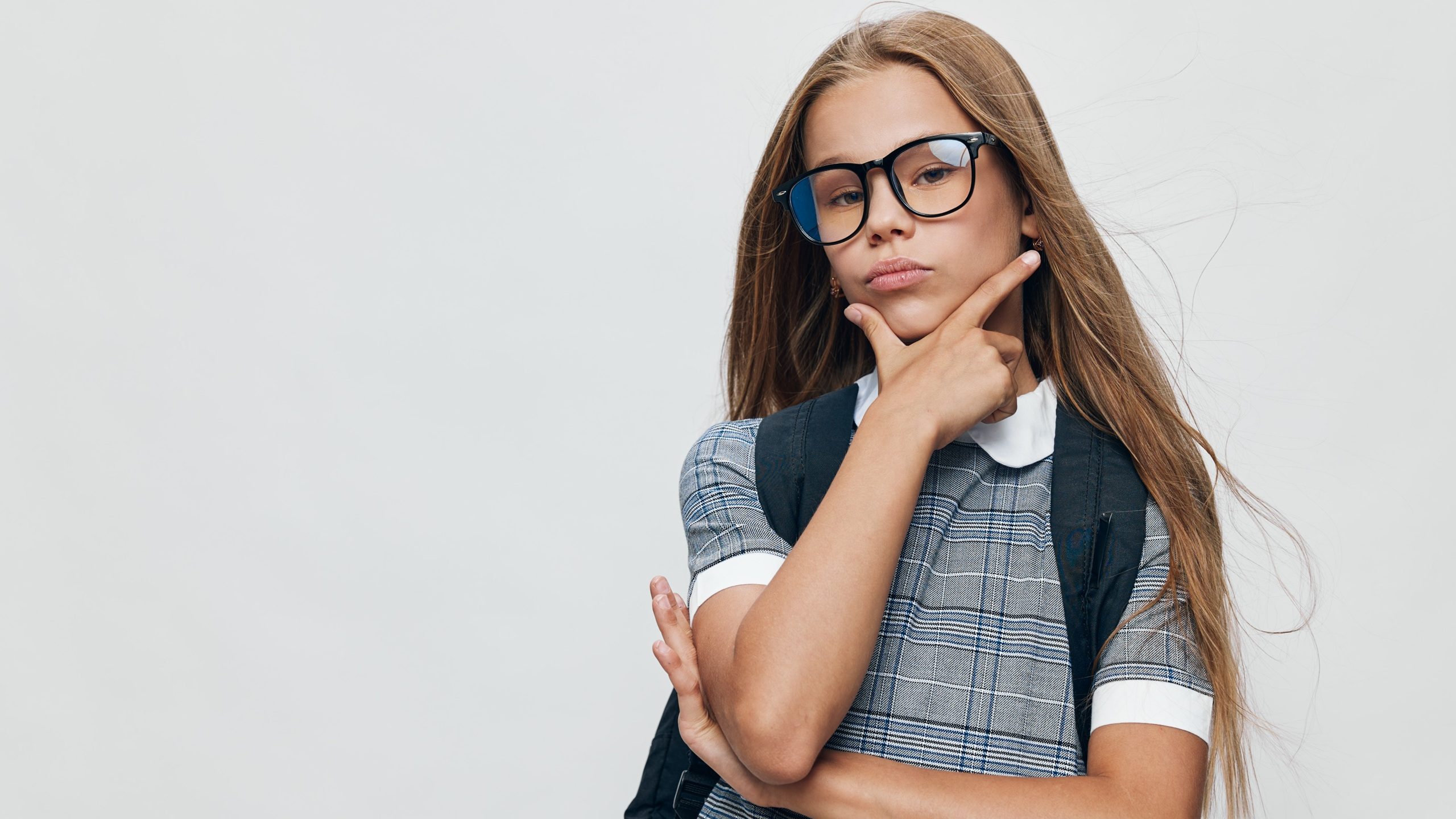 Girl with long hair and glasses wearing a dress with backpack thinking and posing with hand on chin on white background. Youth portrait with casual style and serious expression.