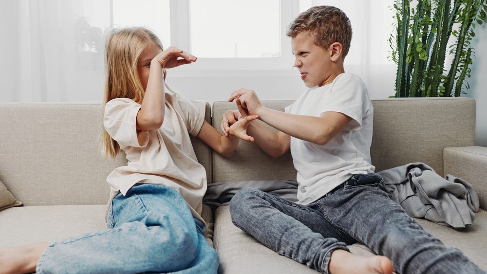 Two children sitting on sofa playfully fighting with expressive faces in bright living room. Concept of sibling rivalry, childhood emotions, and family dynamics.