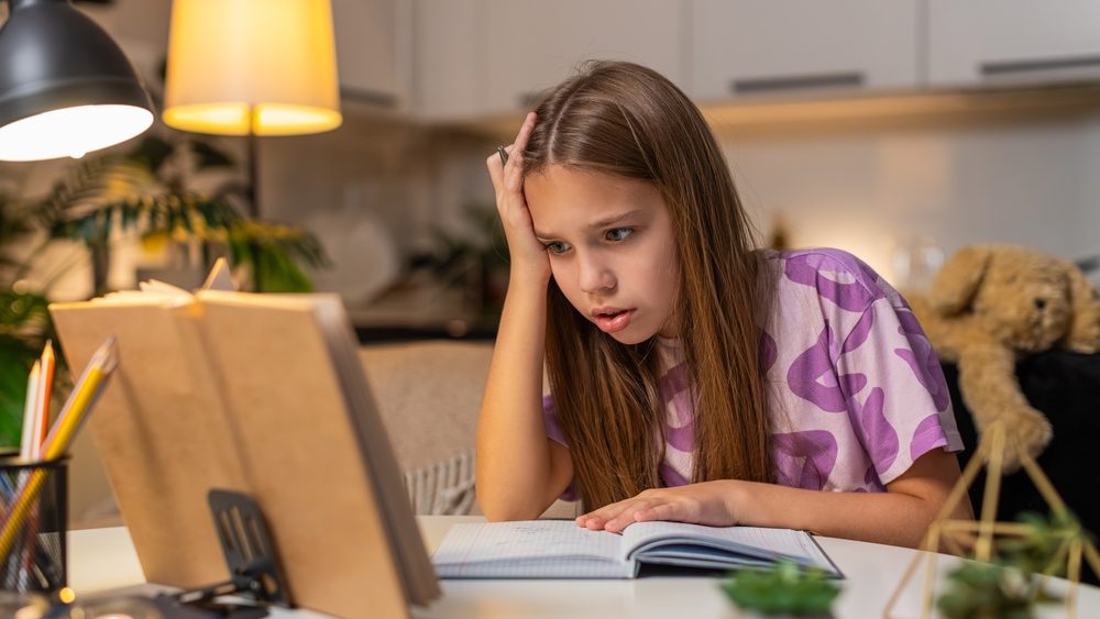 Teenager girl at night home reads problem from book for homework feels confused holds head sighs. Caucasian child kid sitting at table at evening puts notebook aside looks tries to understand slowly.