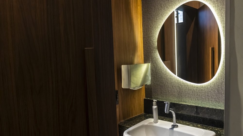 Modern bathroom interior featuring a white ceramic washbasin on a dark granite countertop, an organic-shaped backlit mirror on a textured wall, and warm wooden paneling in a luxury hotel setting