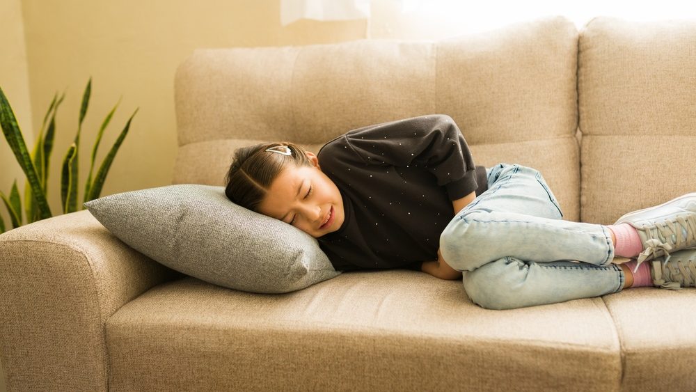 Young girl lying on a couch clutching her abdomen in discomfort, suffering from stomach pain and feeling unwell at home