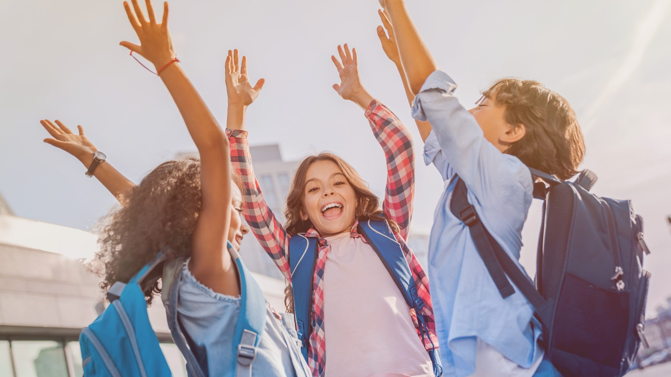 Happy children multiethnic elementary middle school kids pupils classmates friends boys and girls with hands up standing near school outdoors. Expression emotions gestures support unity concept.