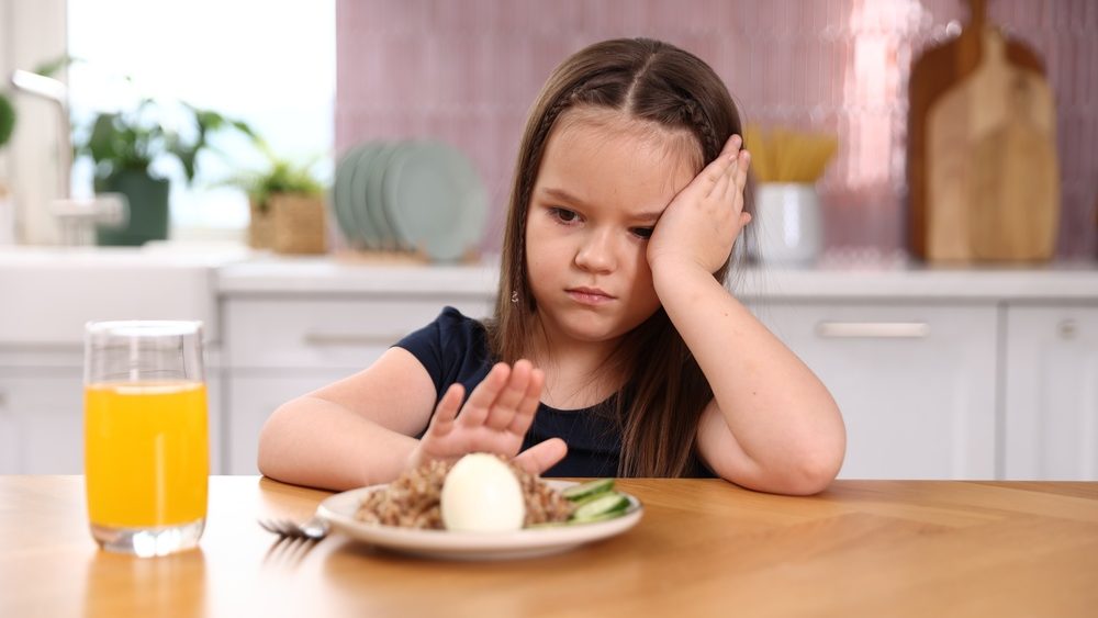 Unhappy little girl refusing to eat buckwheat, egg and vegetables at wooden table in kitchen