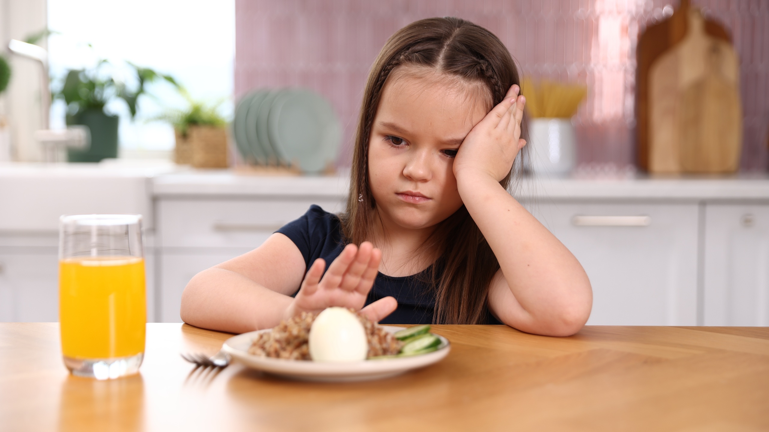 Unhappy little girl refusing to eat buckwheat, egg and vegetables at wooden table in kitchen
