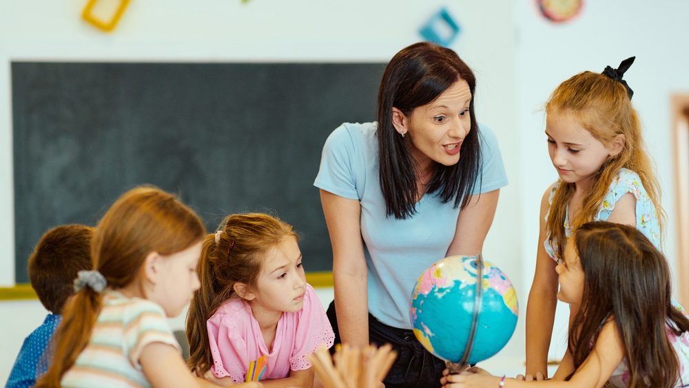 Primary school teacher guiding curious students around a globe during a classroom geography lesson