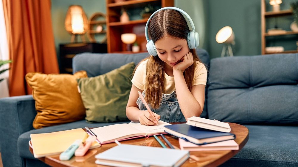 Young girl with headphones studying at a coffee table in a cozy living room, writing in a notebook surrounded by books and stationery.