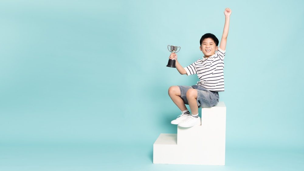 Happy young Asian boy sits on a white pedestal holding a trophy with his arm raised in victory. Success, childhood achievement, and educational excellence