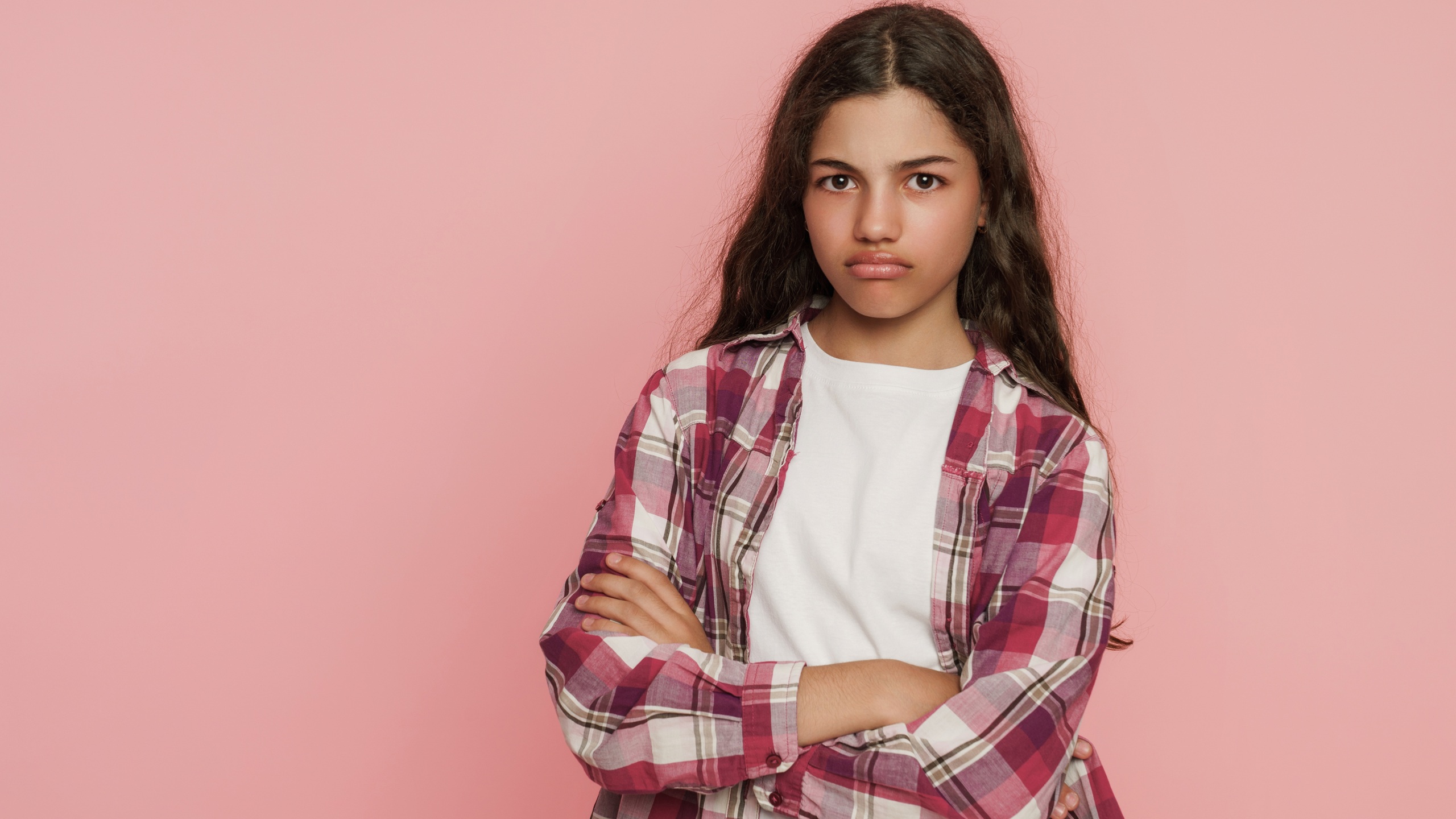 Portrait of displeased mixed race schoolgirl with crossed arms and pouting lips on pink studio background, human emotions, attitude, frustration and childhood expression concept