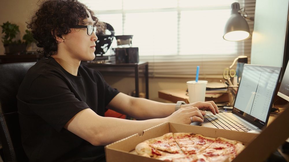 Asian young man working on laptop at home desk with pizza box in foreground, representing remote work lifestyle. Image suitable for food delivery and freelance campaign