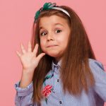 Portrait of funny female kid having long auburn hair looking on camera giving high five, meaning hi or bye with hand over pink background