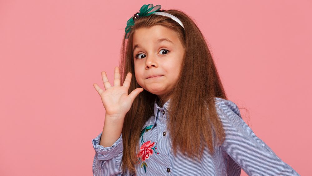 Portrait of funny female kid having long auburn hair looking on camera giving high five, meaning hi or bye with hand over pink background