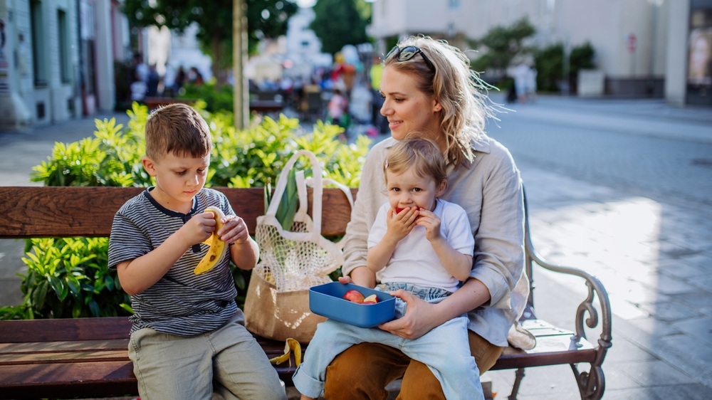 Young mother with little kids resting on bench in city and eating fruit snack.