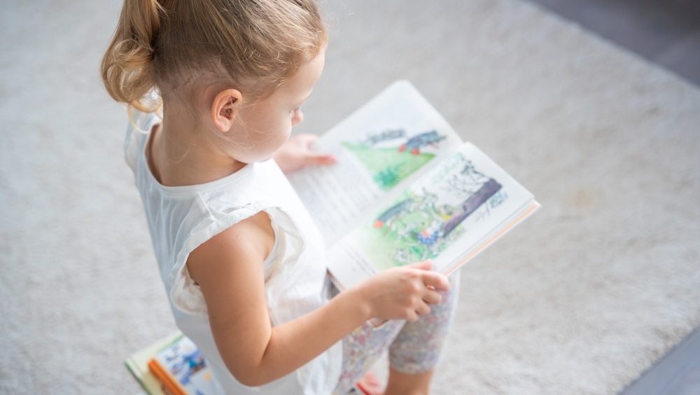 Little girl is sitting on stack of children's books and leafing through a book with pictures of fairy tales. High quality photo