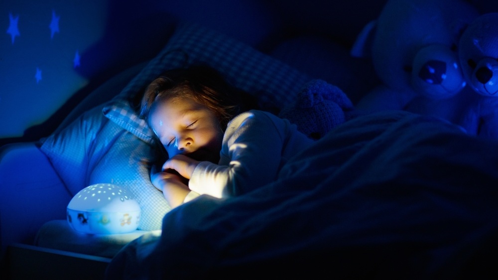 Little girl reading a book in bed. Dark bedroom with night light projecting stars on room ceiling. Kids nursery and bedding. Children read before bedtime. Toddler child playing with lamp and bear toy.