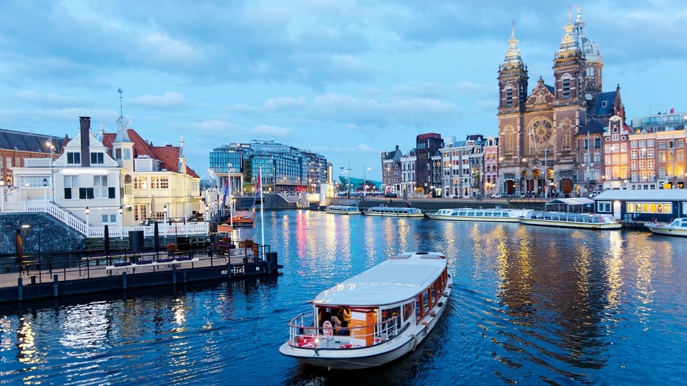 A tour boat cruising on the canal near Amsterdam's Central Railway Station, with the spires of Saint Nicholas Basilica towering in background and city lights reflected in the water at blue dusk
