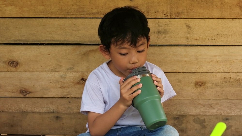 A young Asian boy sips from a green tumbler while seated on a wooden bench indoors with a rustic backdrop.