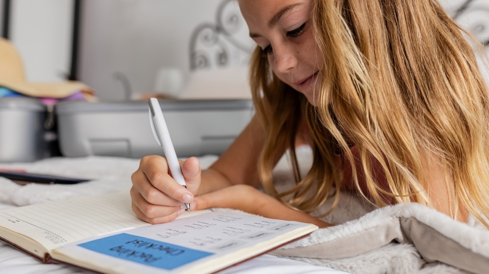 Child writing in notebook on bed at home holding pen near suitcase with straw hat. Journaling, studying, packing, anticipation, concentration, creativity, cozy