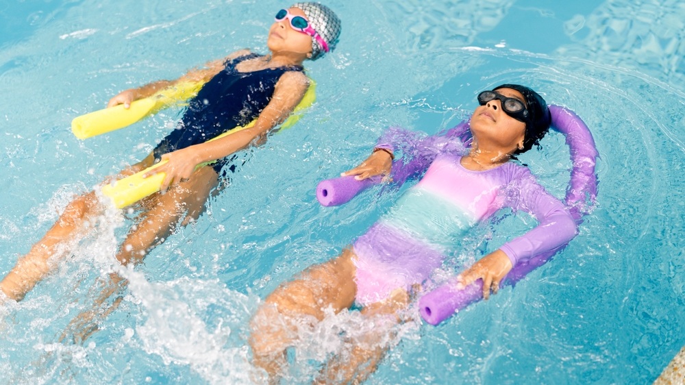 Two young girls practicing backstroke and floating independently with pool noodles in a bright blue swimming pool, splashing water