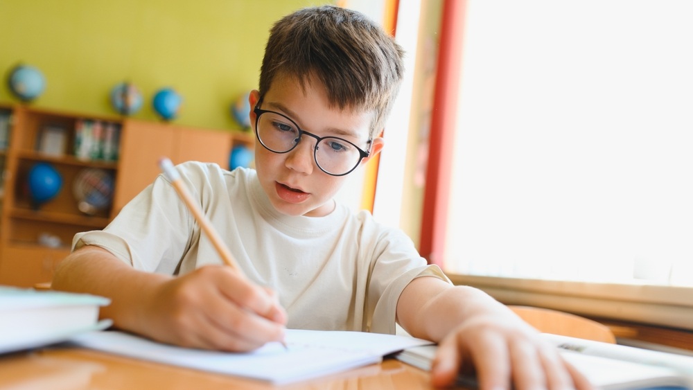 Young student wearing eyeglasses writing on notebook with pencil, studying at desk in classroom