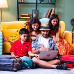 Happy Indian family of four with laptop and suitcases in modern home doing web check-in or booking an international holiday trip together in summer attire