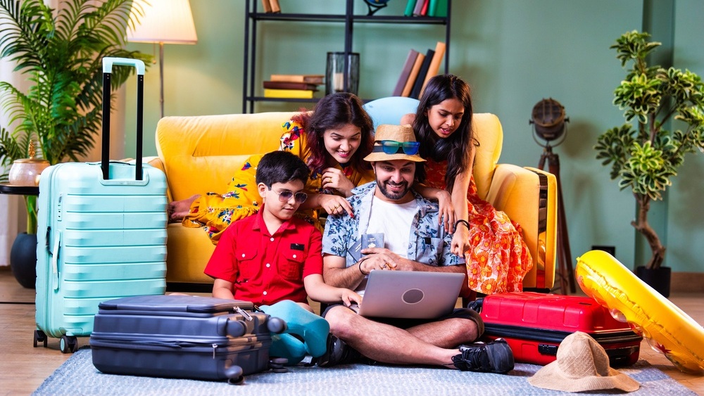 Happy Indian family of four with laptop and suitcases in modern home doing web check-in or booking an international holiday trip together in summer attire