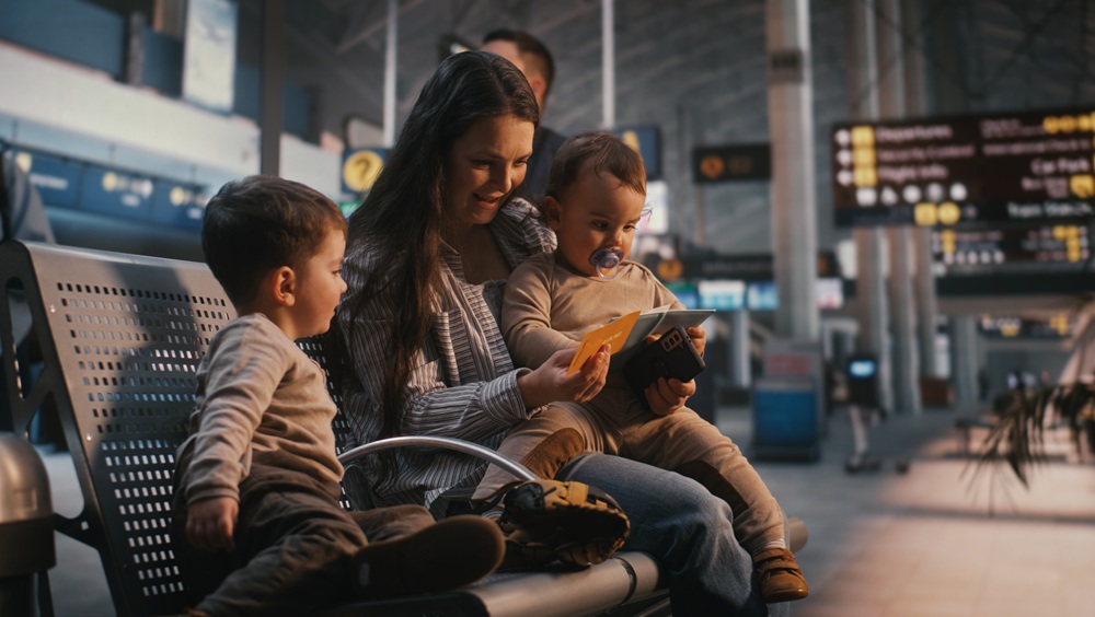 Young Mother Sits in Departure Lounge, Holding Baby and Showing Travel Documents to Older Son. Family Travel, Parenting Challenges During Trips, Anticipation Holiday Flight at International Airport.