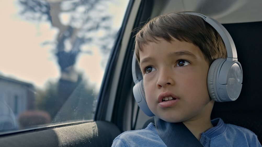 A young boy sits in a car, wearing headphones, gazing out the window. He appears curious and engaged, perhaps listening to music or an audiobook.