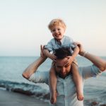 Father with a toddler boy standing on beach on summer holiday, having fun.