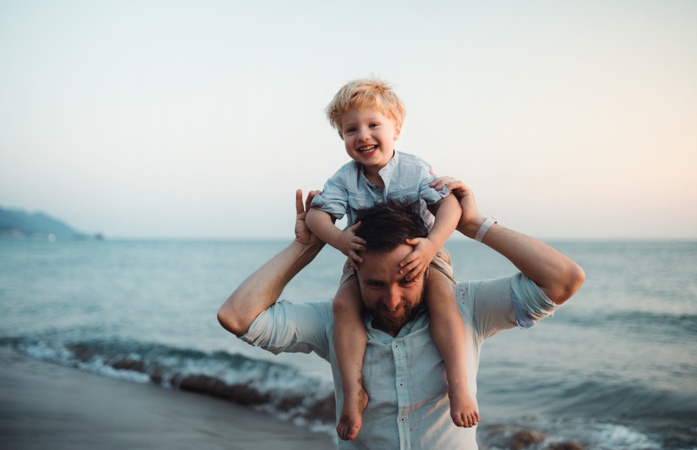 Father with a toddler boy standing on beach on summer holiday, having fun.