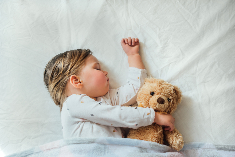 Toddler baby sleeping on white sheets hugging soft brown teddy bear. Children's nap time or bedtime. Child sleep deprivation. Child sleep routine and schedule.