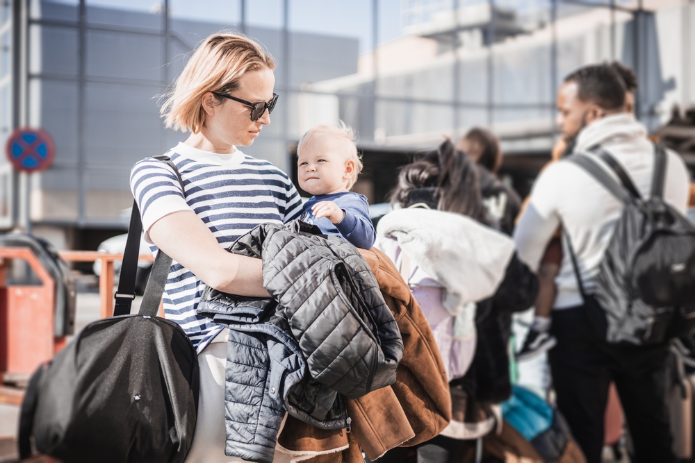 Motherat travelling with his infant baby boy child. Mom holding travel bag and her infant baby boy child while queuing for bus in front of airport terminal station.
