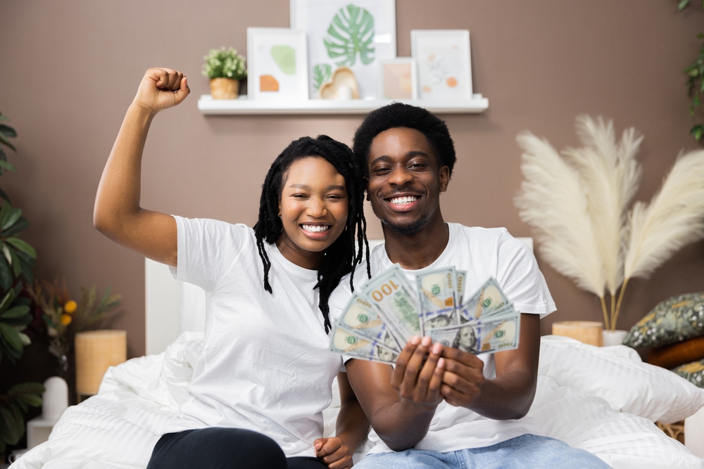 Smiled young African American dark skinned couple man and woman wearing casual white t-shirt smiling raising fists holding a lot of money cash in hands celebrating happi lottery winners.