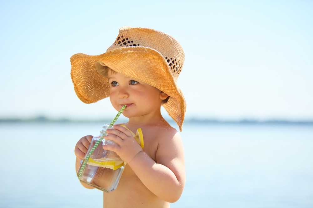 Child in straw hat drinking lemonade with straw on beach. Concept of summer vacation, refreshment, innocence, childhood joy, health, hydration and leisure.
